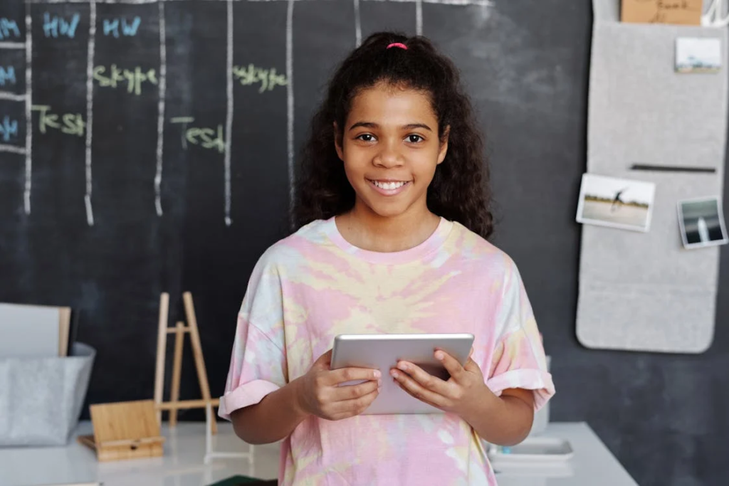 A young girl smiling with a tablet in front of a chalkboard